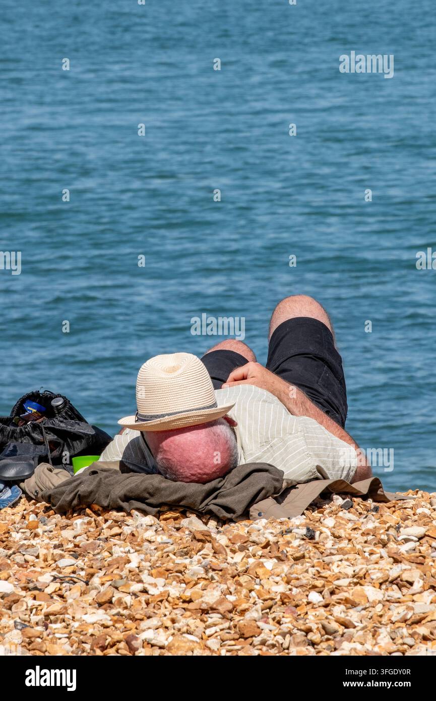 Uomo anziano con scottature rosse sulla sommità della testa e un cappello che copre il viso sdraiato al sole su una spiaggia di ciottoli a Cowes sull'isola di Wight, Regno Unito Foto Stock