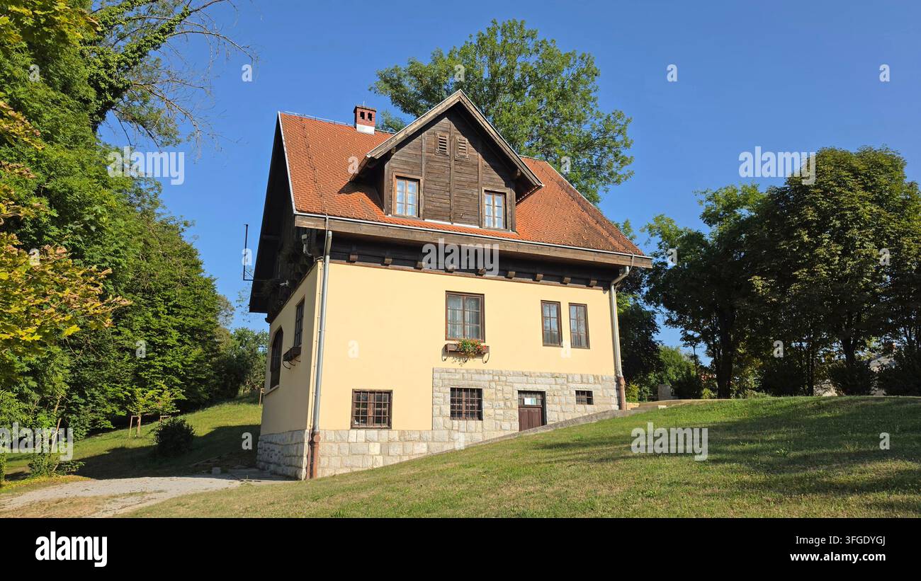 Casa di campagna rustica con fondazione in pietra, sottotetto in legno e tetto di tegole rosse, situata di fronte a una fitta foresta verde su un pendio erboso sotto un cielo blu terso Foto Stock