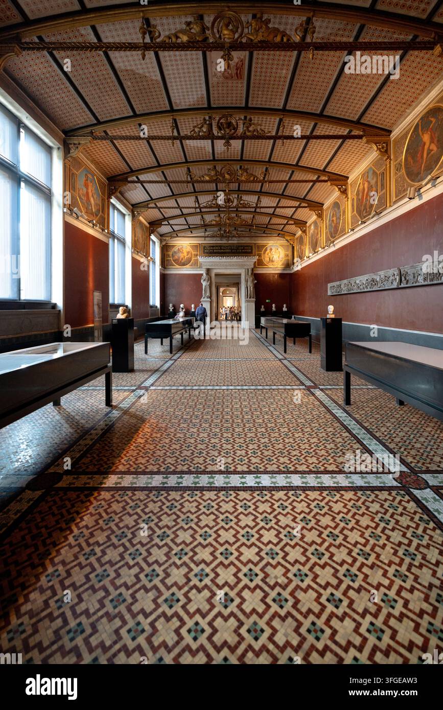 I visitatori esplorano reperti archeologici in una lunga galleria a volta del Neues Museum, dove archi e colonne in mattoni restaurati rivelano la struttura dell'edificio Foto Stock