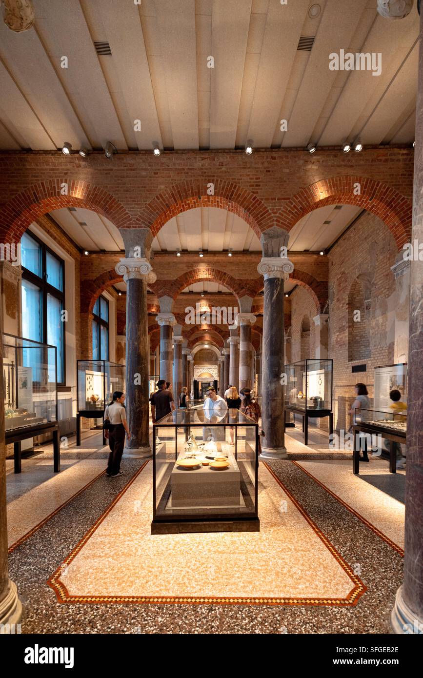 I visitatori esplorano reperti archeologici in una lunga galleria a volta del Neues Museum, dove archi e colonne in mattoni restaurati rivelano la struttura dell'edificio Foto Stock