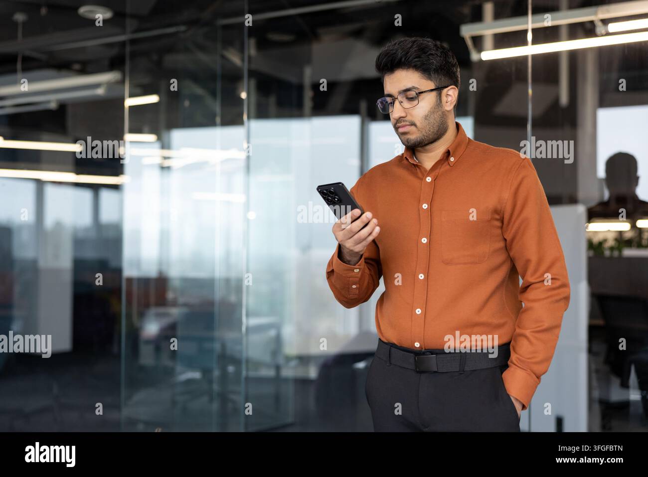 Ritratto di un giovane professionista in abito da lavoro in piedi in un ufficio contemporaneo in vetro, che controlla con calma il suo smartphone vicino a una parete di vetro con riflessi sottili e illuminazione interna soffusa Foto Stock