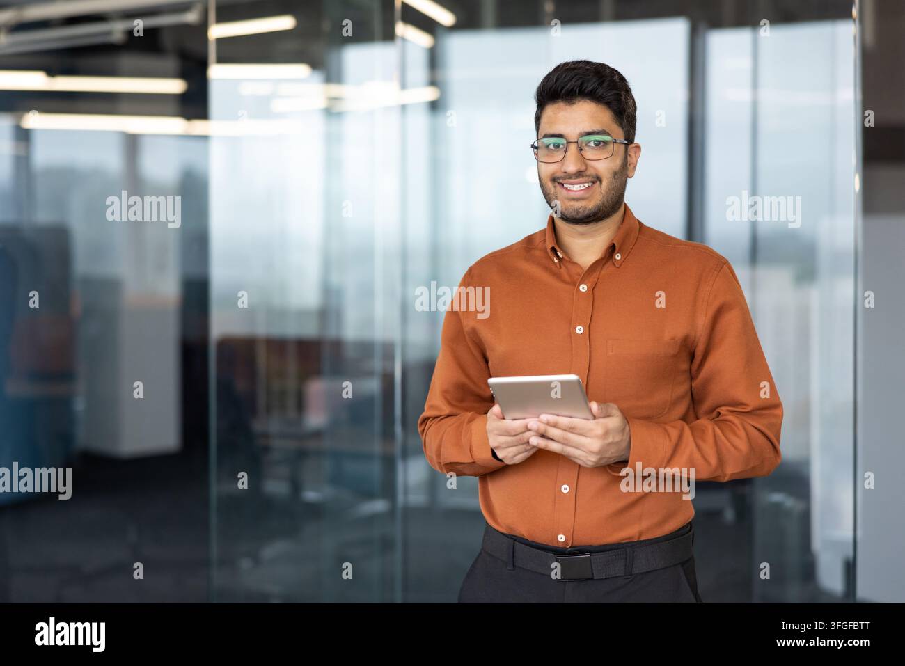Ritratto di un giovane professionista sorridente mentre tiene un tablet in un luminoso corridoio di ufficio contemporaneo, con pareti di vetro, riflessi morbidi e luci a soffitto, con uomo, sorridente, vetro, dettaglio del tablet Foto Stock