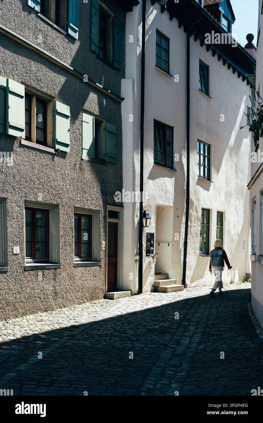 Vicolo stretto baciato dal sole in una vecchia città idilliaca con ombre profonde sulle facciate storiche e una passeggiatrice solitaria sullo sfondo, Knoepfle Foto Stock
