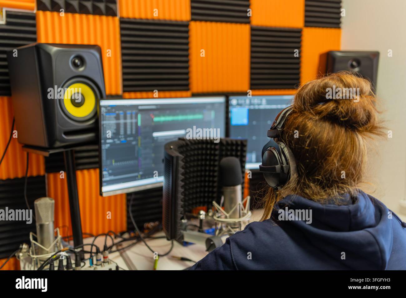 Donna con cuffie sopra l'orecchio modifica l'audio su software DAW su due monitori, affiancata da altoparlanti da studio e microfoni a condensatore in aco Foto Stock