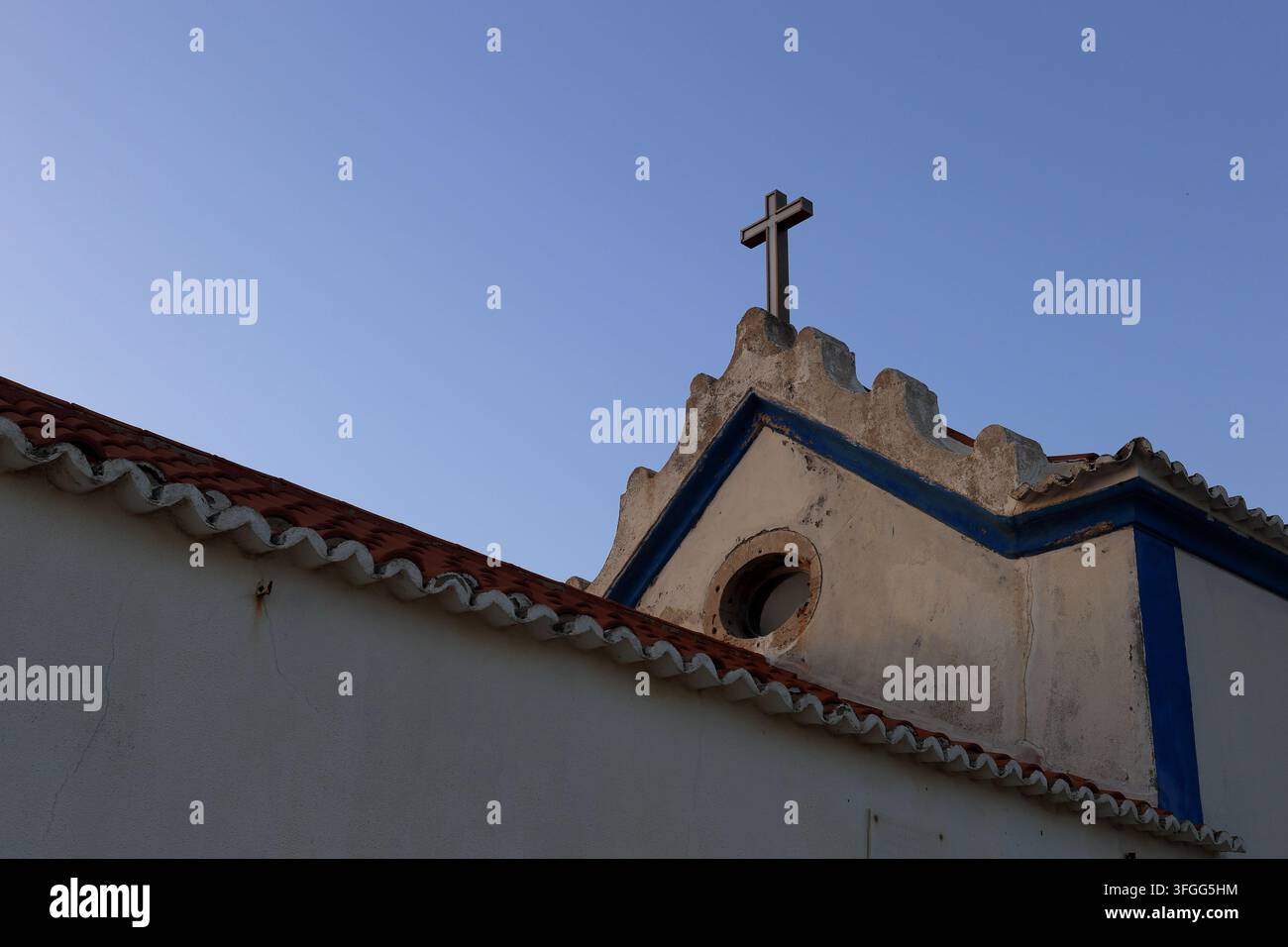 Barril, Portogallo - 03 settembre 2026: Cappella color crema consumata con rifiniture azzurre, cornici ornate sul tetto, croce in legno, finestra circolare e decorativa Foto Stock