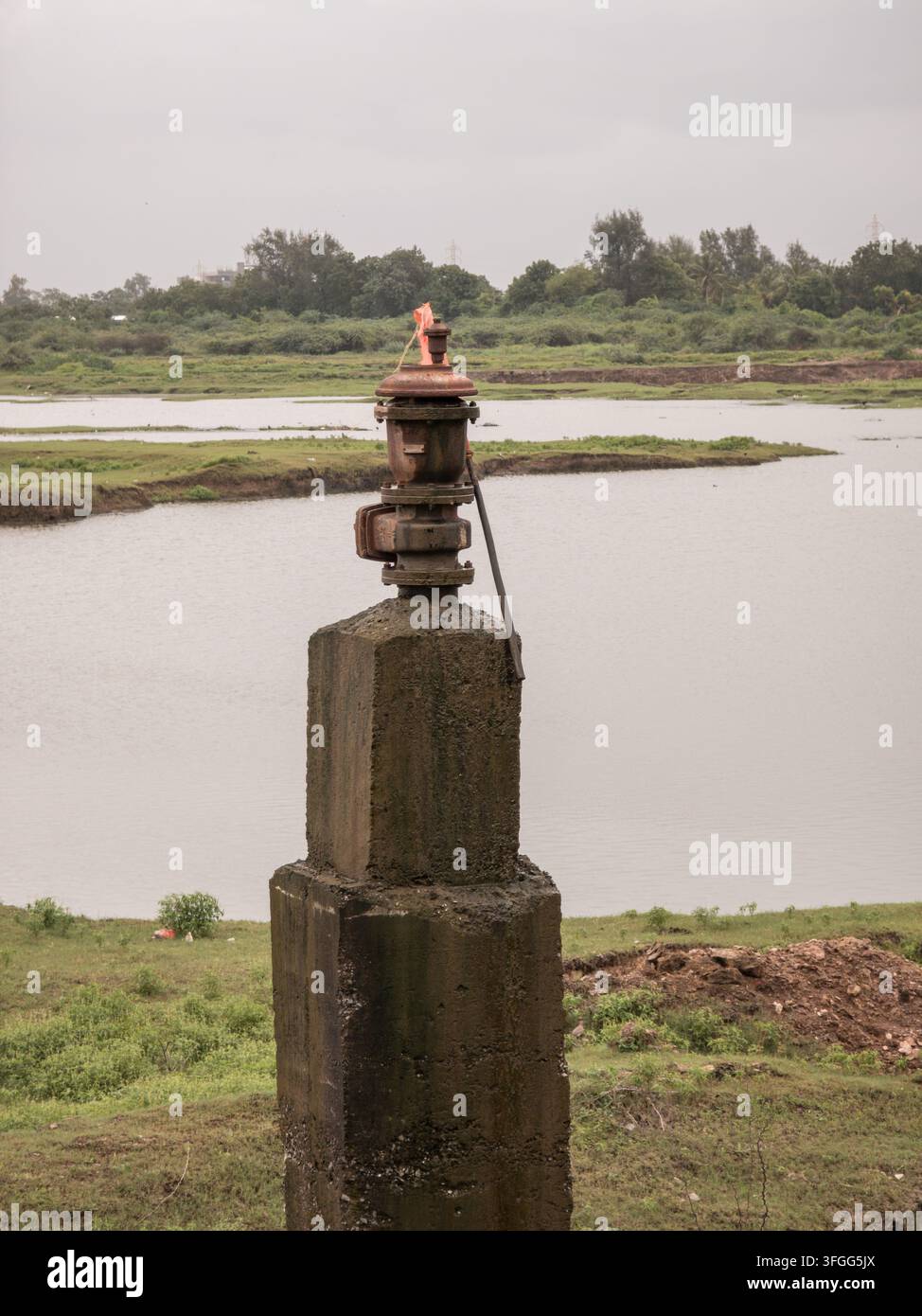 Una valvola dell'acqua consumata si erge in modo prominente su un pilastro di cemento accanto a un fiume sereno nel Gujarat, India, circondata da una vegetazione lussureggiante e cieli nuvolosi - Immagine stock catturata con smartphone