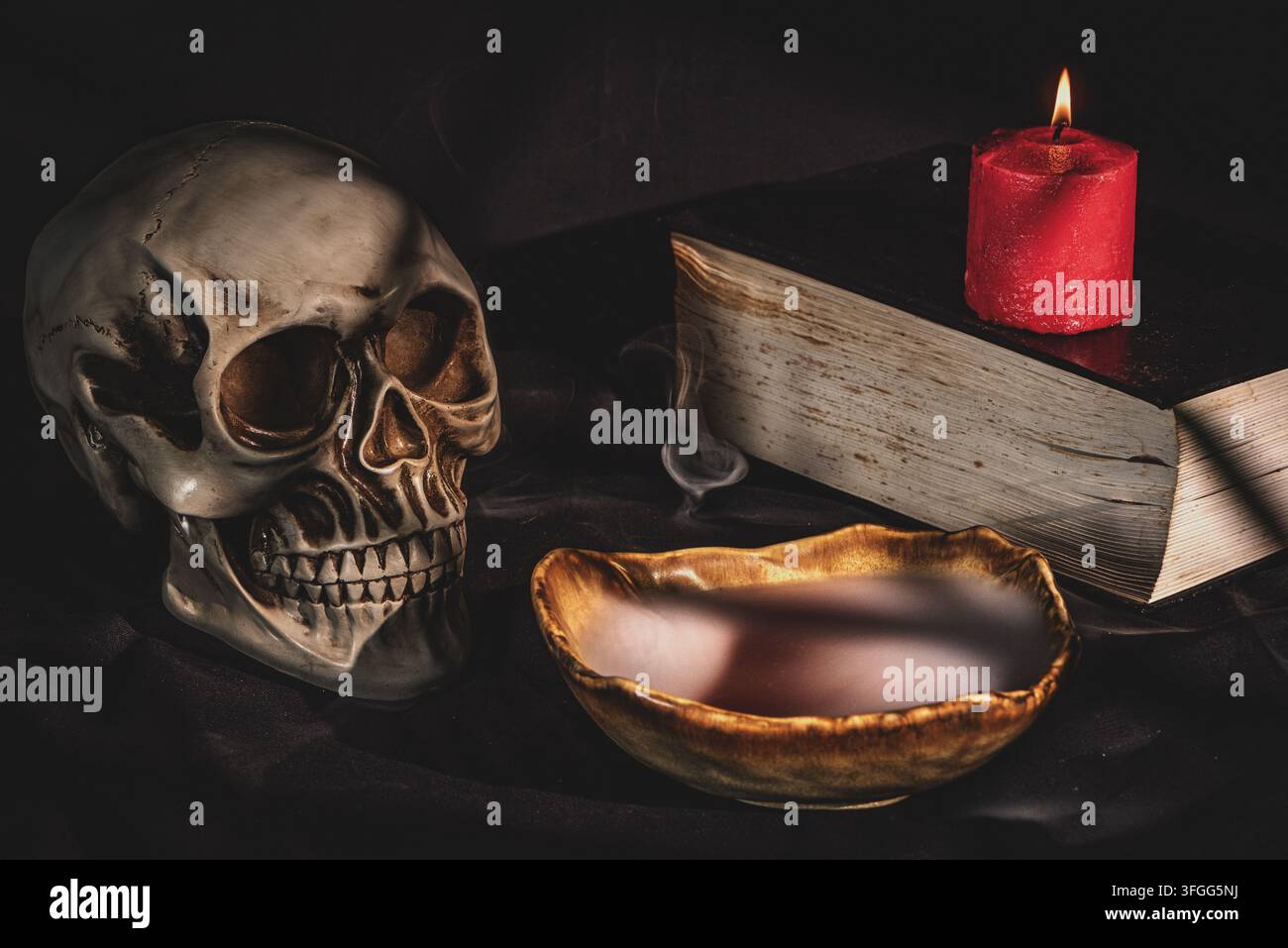 Teschio umano con candela accesa, libro antico e misteriosa pozione rossa in una ciotola fumante Foto Stock