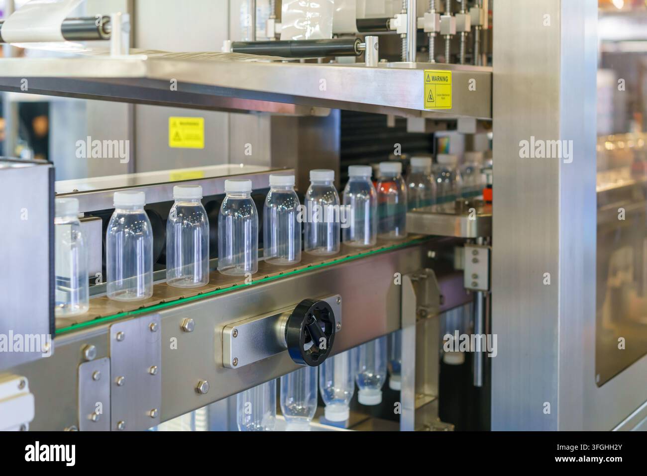 Bottiglie di plastica trasparenti vuote con tappi bianchi che avanzano su un nastro trasportatore in acciaio inossidabile all'interno di uno stabilimento di produzione industriale automatizzato. Foto Stock