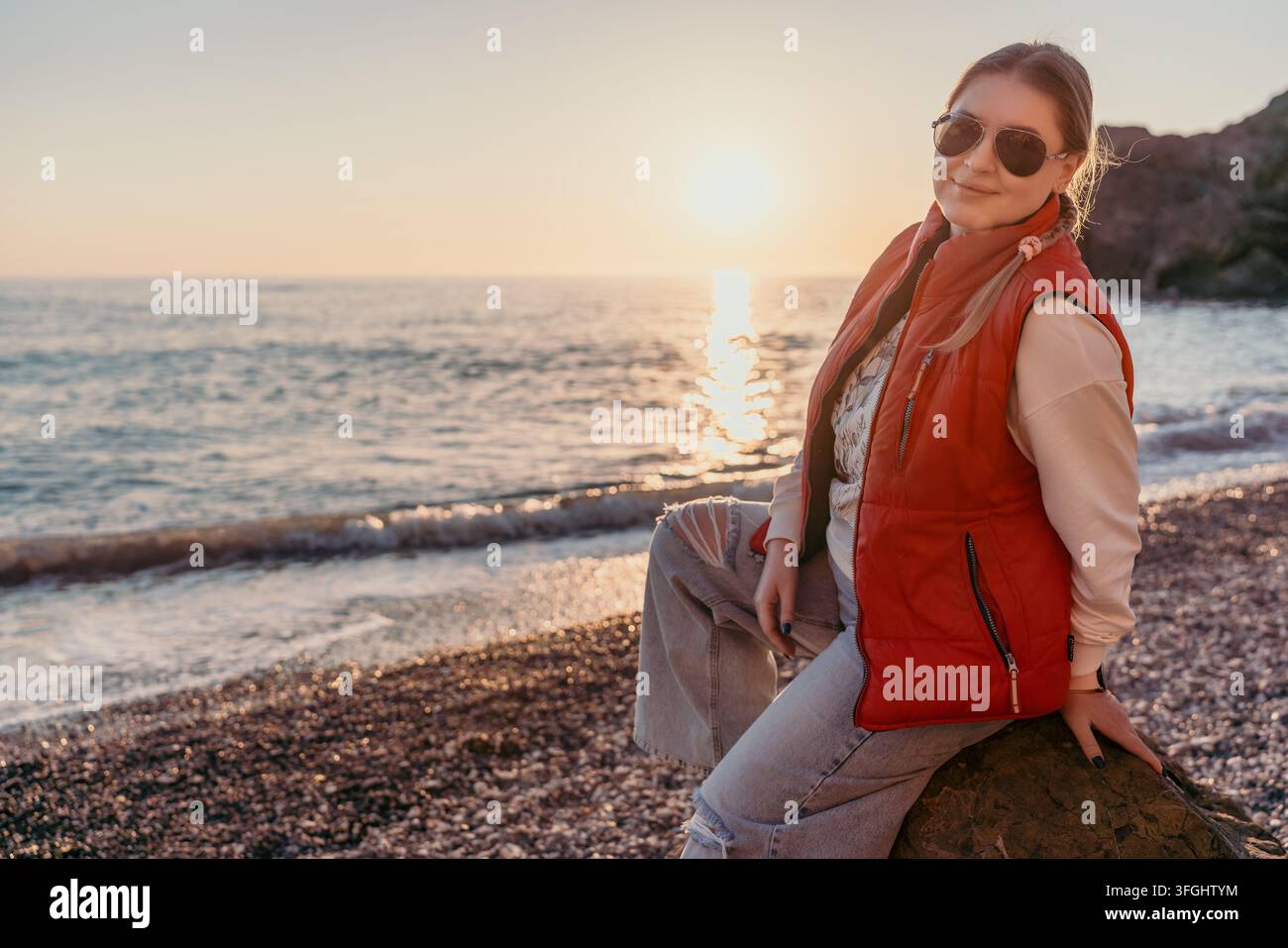 Donna spiaggia tramonto giovane donna in un gilet rosso seduta su una riva di ciottoli che gode di una vivace vista sull'oceano dell'ora dorata Foto Stock