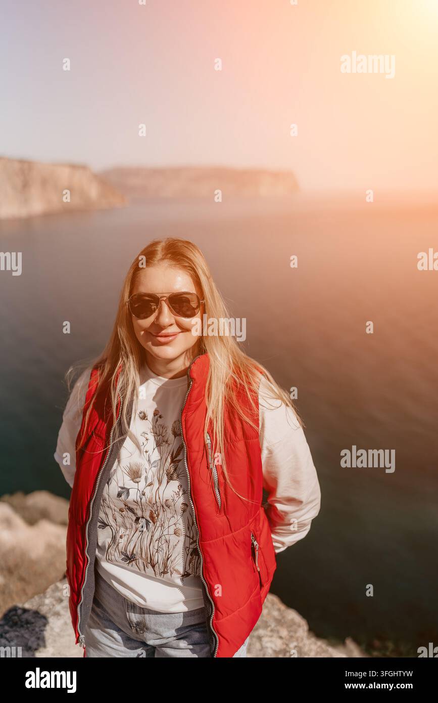 Donna viaggio costiero, signora bionda sorridente con un giubbotto rosso in piedi su una scogliera rocciosa che si affaccia sul mare durante una giornata soleggiata Foto Stock