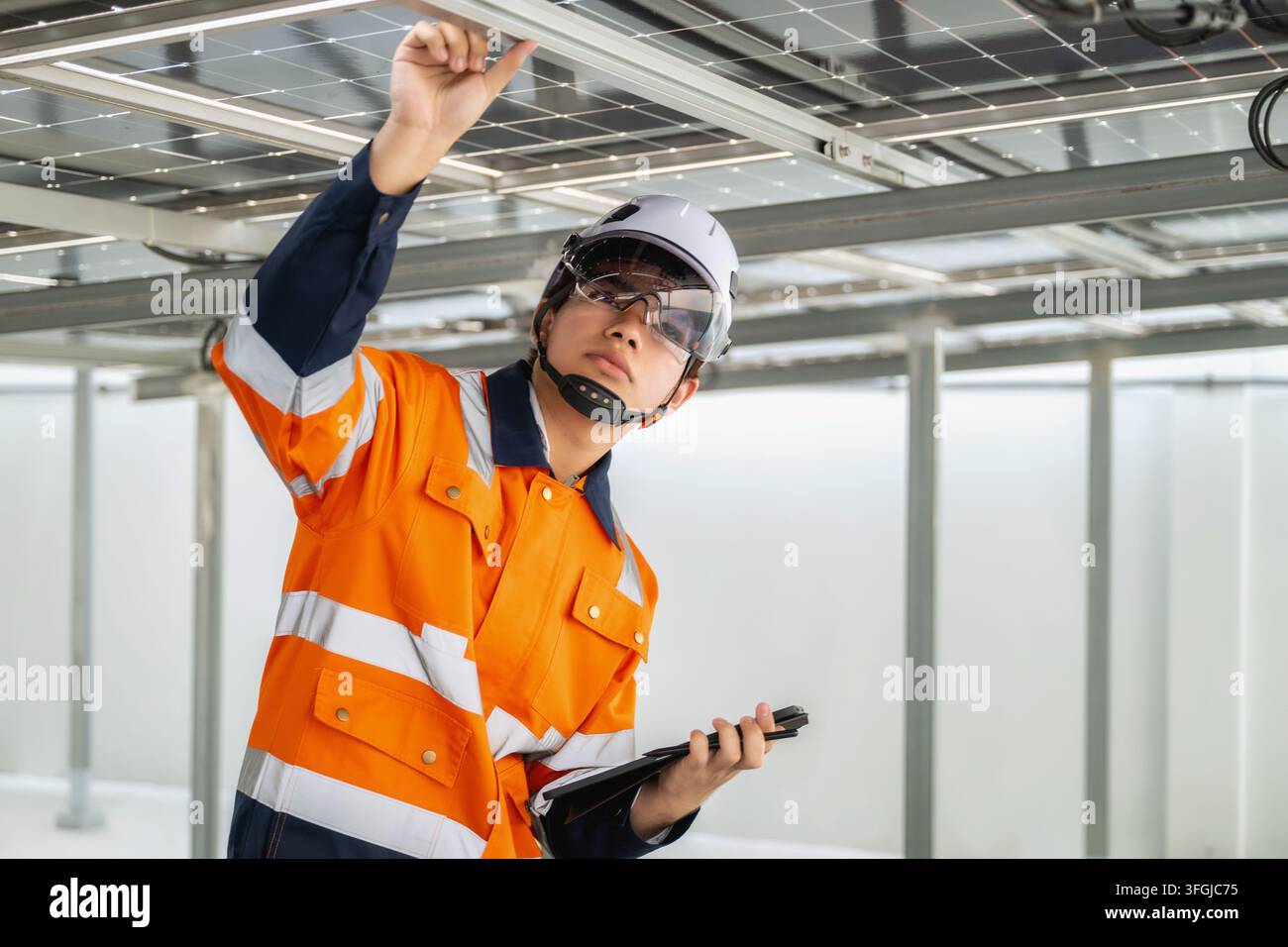 Giovane ingegnere in uniforme arancione e casco bianco che esamina pannelli solari e strutture a travi metalliche, rivedendo i dettagli dell'installazione su un tablet. espressione Foto Stock
