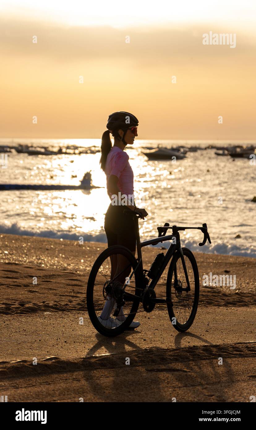 Silhouette di profilo di una ciclista che guarda felice il mare, in piedi accanto alla sua bici da corsa sulla spiaggia durante l'ora dorata al tramonto a Toss Foto Stock