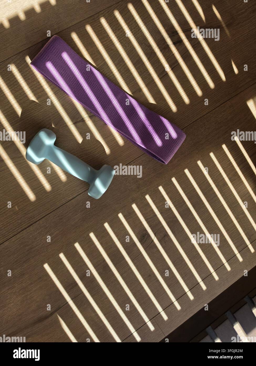 Manubrio blu chiaro e fascia di resistenza viola su un pavimento di legno con persiane che creano un motivo di luce e ombra - Immagine stock catturata con smartphone