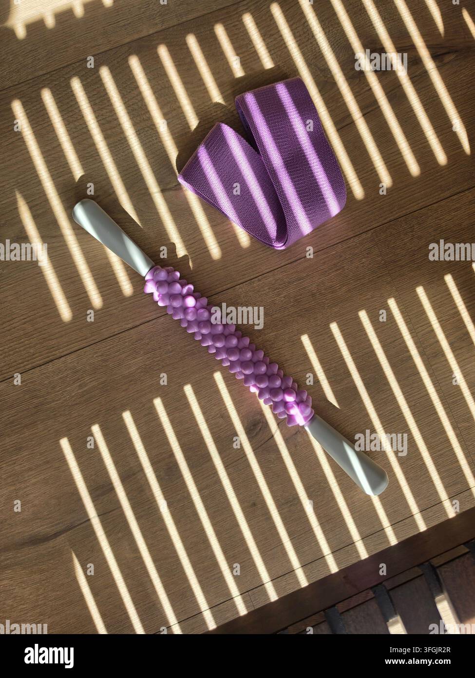 Rullo da massaggio ruvido e fascia di resistenza piegata su un pavimento di legno con persiane che creano un motivo di luce e ombra - Immagine stock catturata con smartphone