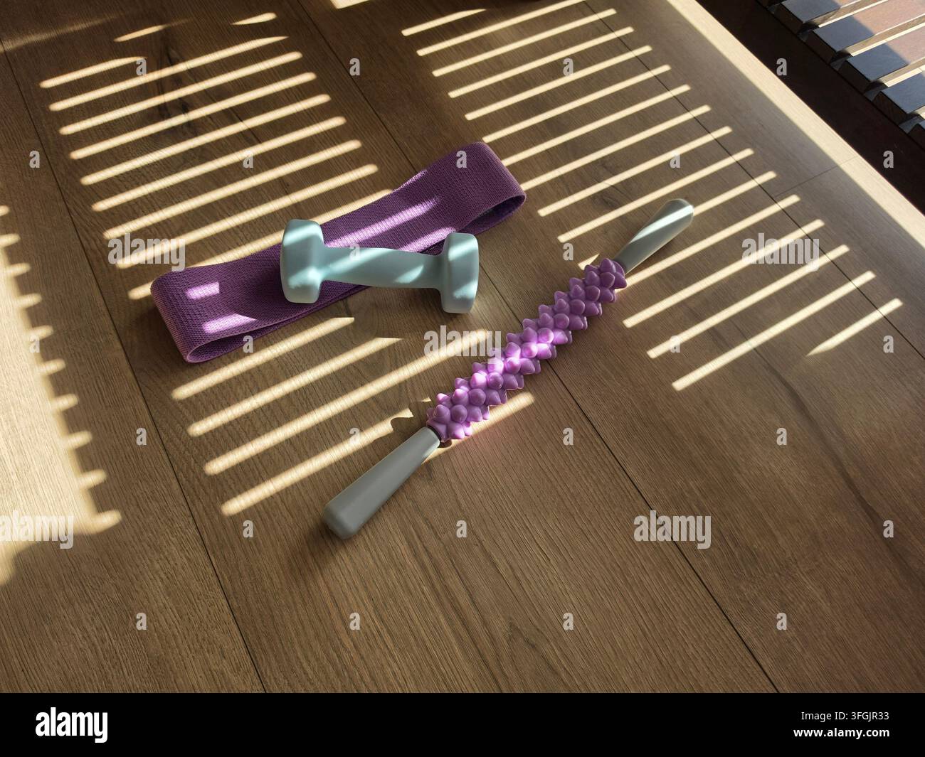 Accessori per il fitness tra cui fascia di resistenza, piccolo manubrio e rullo massaggiante su un pavimento di legno con persiane che creano un motivo di luce e ombra Foto Stock