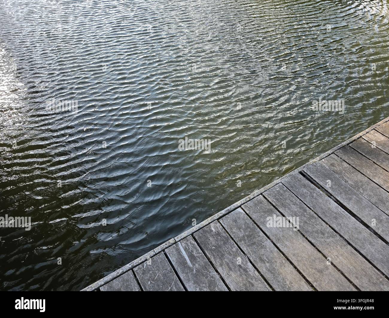 Vista ravvicinata della superficie dell'acqua tranquilla con delicate increspature accanto a un molo di legno sul bordo del pontile in una luminosa giornata di sole - Immagine stock catturata con smartphone