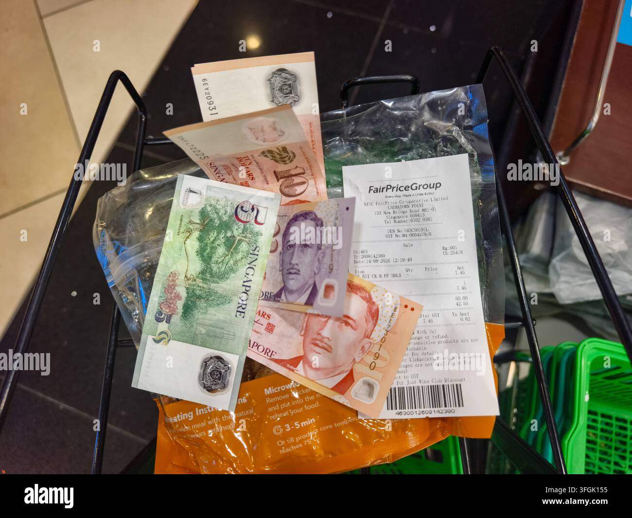 Banconote del Singapore Dollar (SGD) insieme a una ricevuta del supermercato FairPrice Group posizionata dentro un cestino della spesa Foto Stock