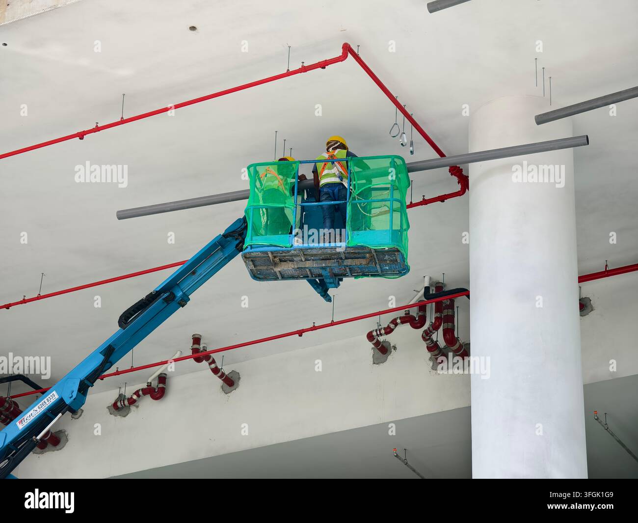 Operai edili con giubbotti ad alta visibilità e caschi gialli all'interno della gabbia della piattaforma verde di un sollevatore a braccio articolato, Singapore Foto Stock
