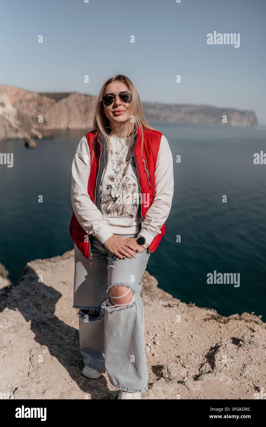 Donna viaggiatrice costiera in piedi su una scogliera rocciosa che si affaccia su un oceano blu sotto un cielo sereno durante un viaggio avventuroso Foto Stock
