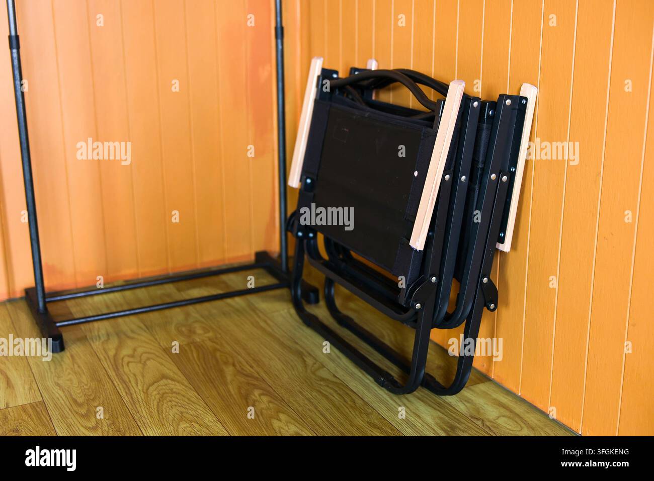 Una sedia pieghevole in metallo nero appoggiata a un muro di legno arancione in un ambiente interno con pavimento in legno lucido Foto Stock