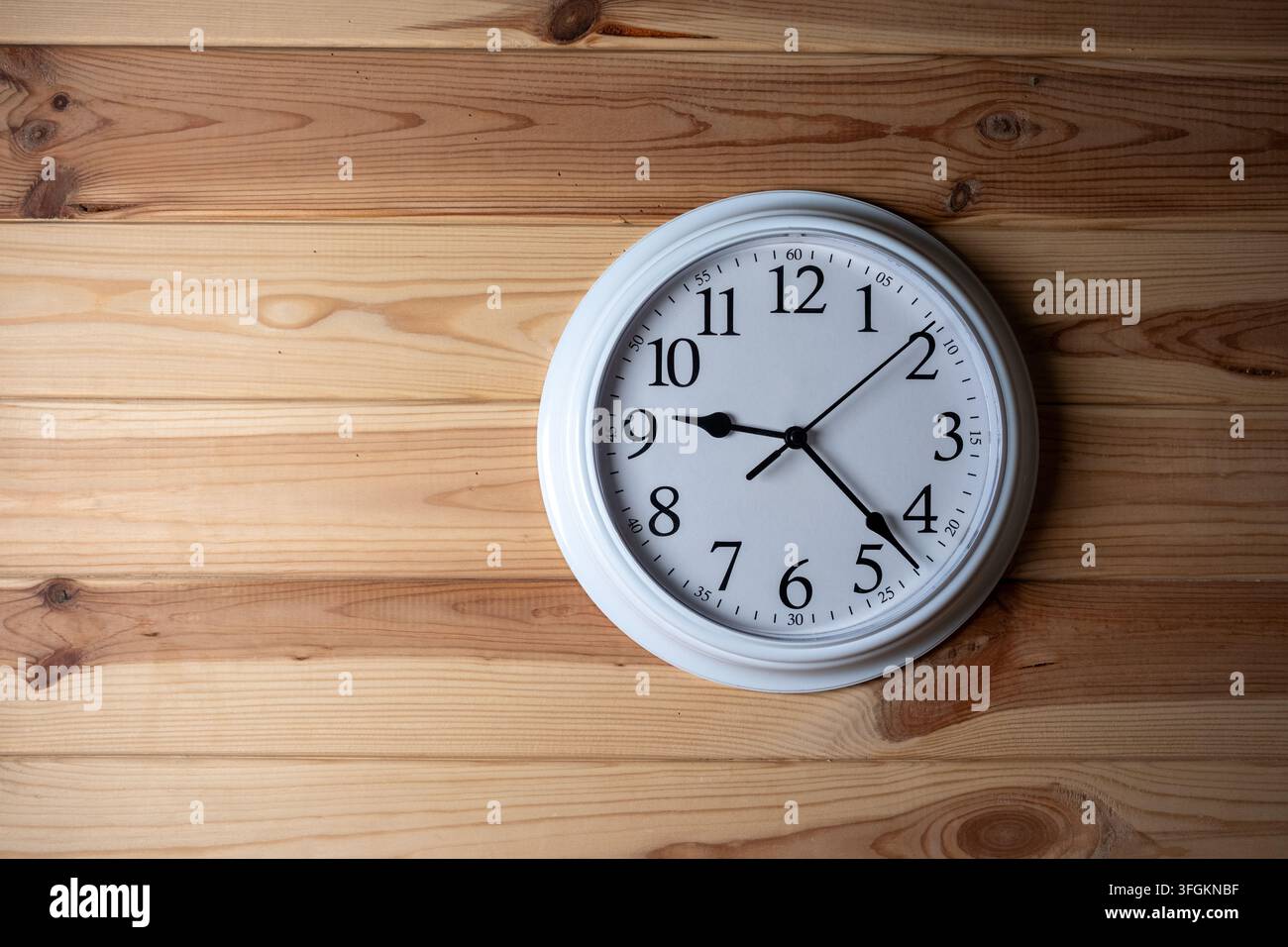 Un orologio da parete analogico bianco rotondo con frecce nere appeso su uno sfondo di legno leggero texturizzato con spazio per il testo Foto Stock