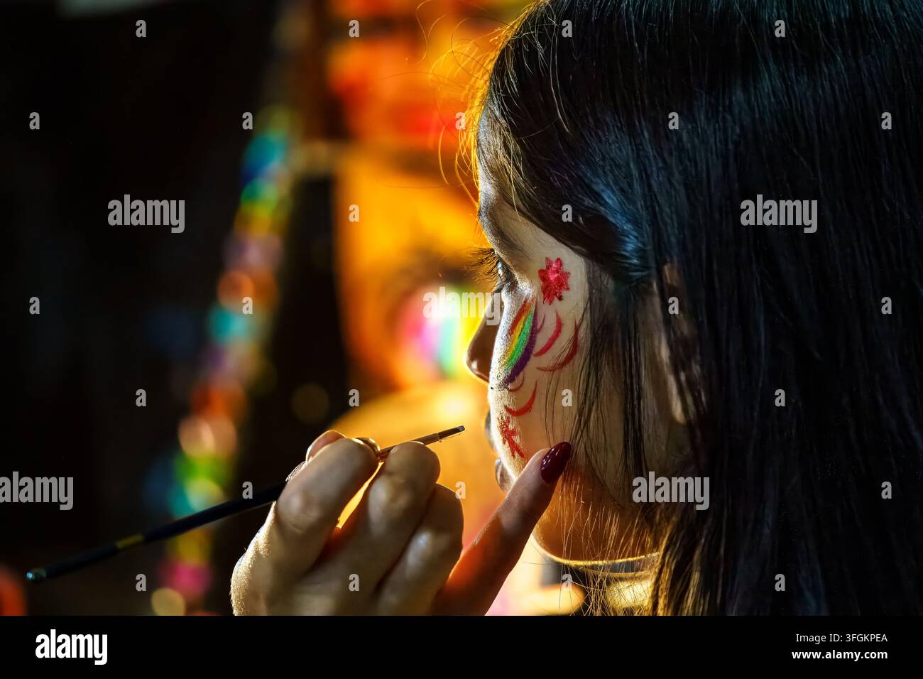 Una luce soffusa illumina il suo viso. Pittura colorata adorna la sua guancia con cura gentile. Il suo sguardo mostra una concentrazione quieta. Il pennello si immerge in tonalità vibranti. Perfe Foto Stock