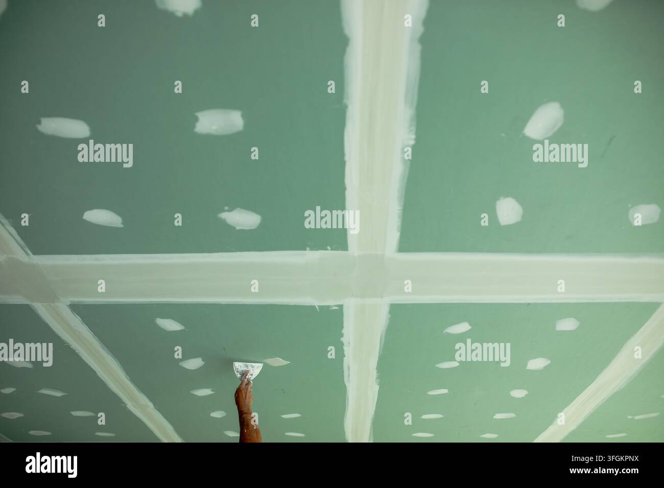 Lavoratore si protende verso l'alto con la spatola, applicando la malta per giunti su una giuntura sigillata con nastro adesivo su un soffitto in cartongesso verde, giunti incrociati e macchie sparse segnano ong Foto Stock