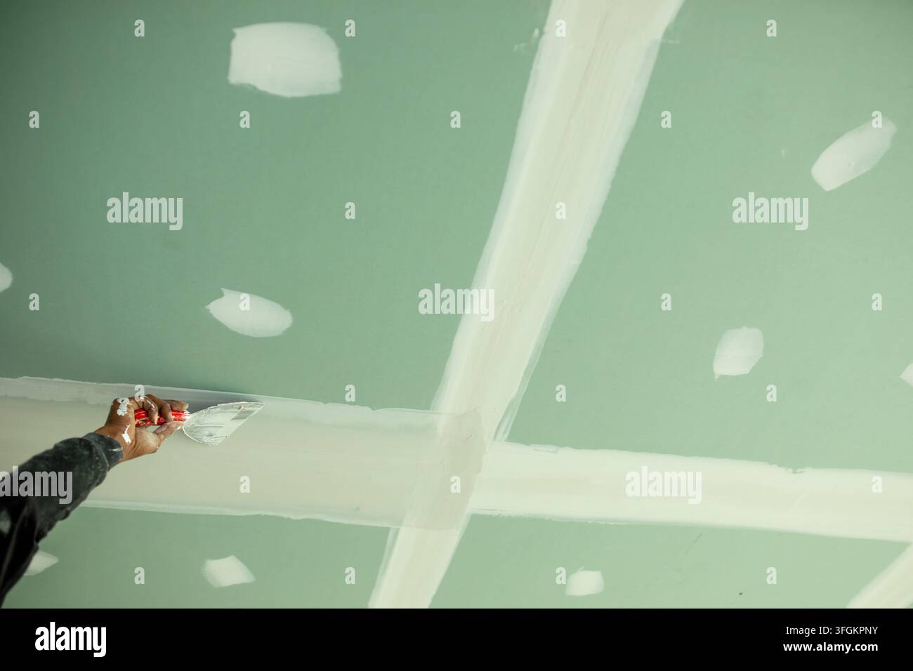 Mano di un operaio tiene una spatola per lisciare la malta lungo una giuntura di cartongesso sigillata, il soffitto in cartongesso verde mostra giunture incrociate e una vite riparata Foto Stock