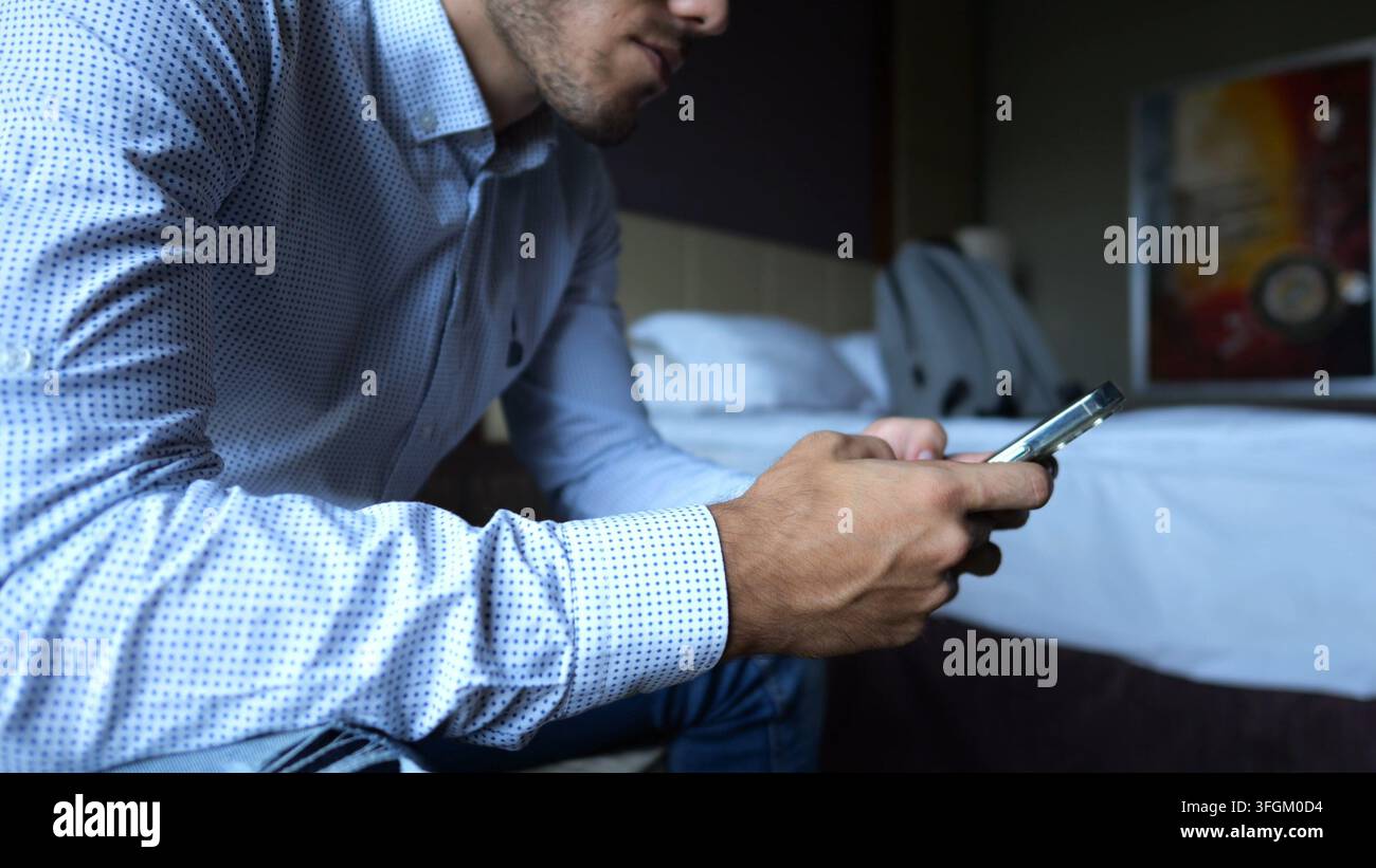 Imprenditore maschile che naviga sul moderno smartphone seduto in una moderna camera d'hotel o in un appartamento. Uomo d'affari di successo che utilizza la tecnologia mobile Foto Stock