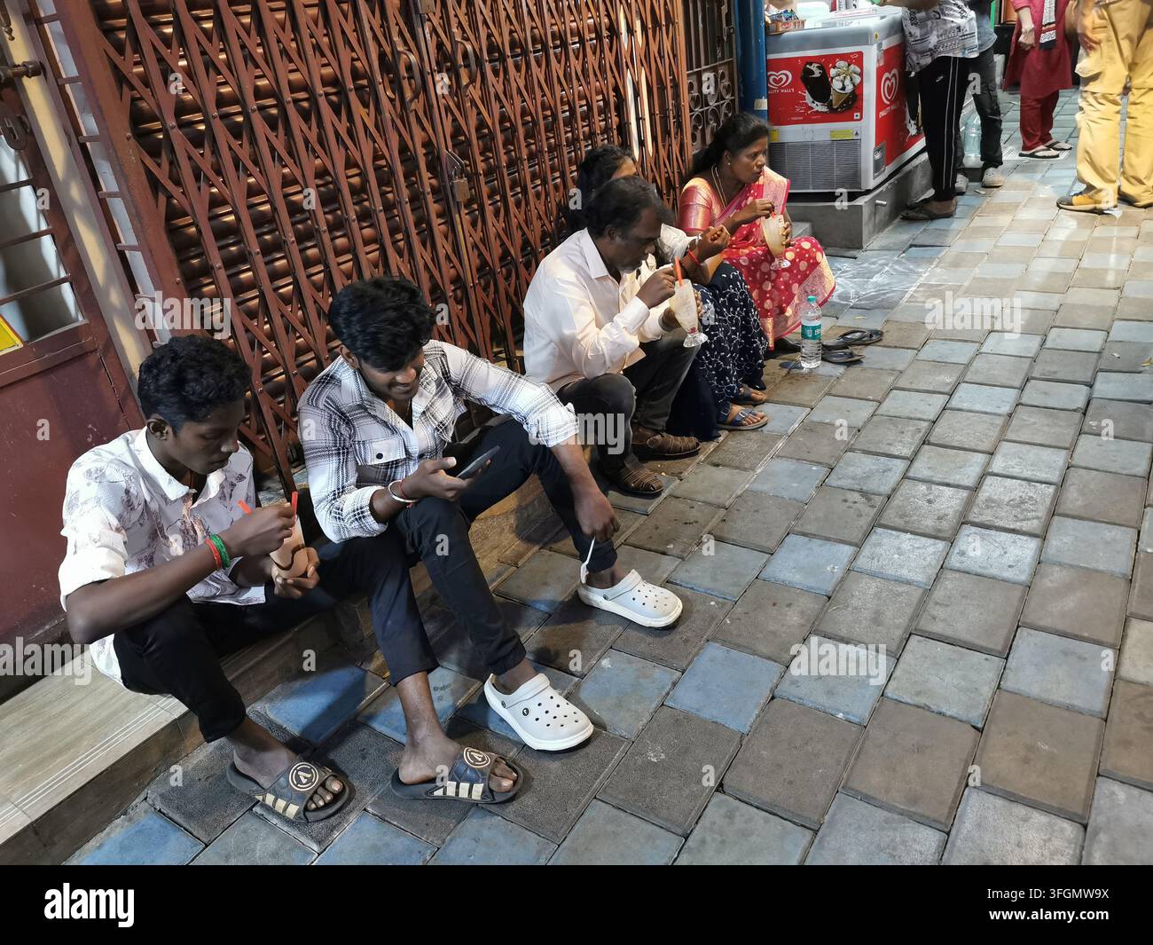 PONDICHERRI, INDIA - 17 GENNAIO 2026: Un gruppo di persone, tra cui due giovani uomini assorbiti dai loro smartphone, seduti sui gradini di pietra mentre bevono Foto Stock