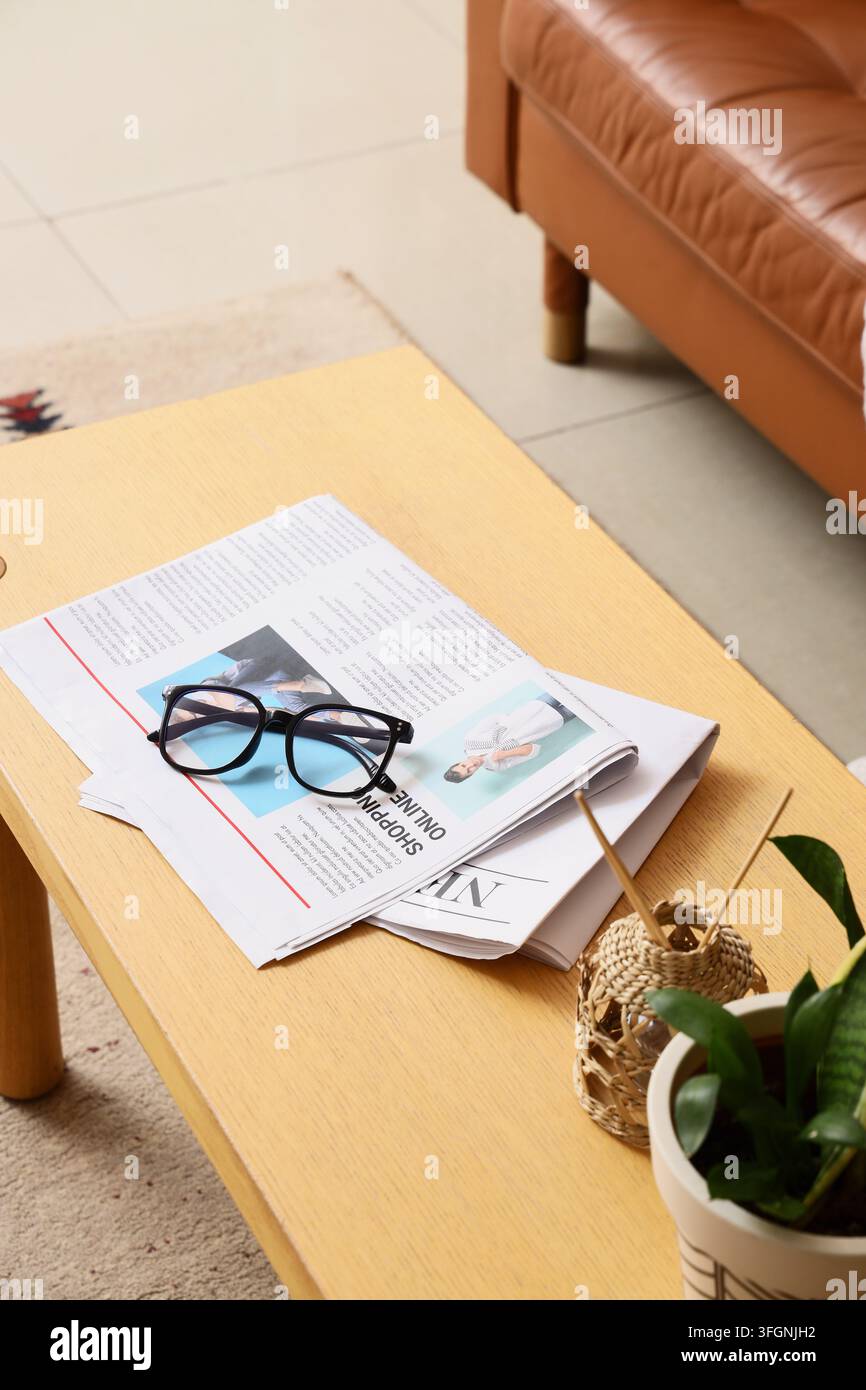 Shopping online e tendenze dell'e-commerce, occhiali appoggiati su un giornale su un tavolo di legno in un accogliente interno di soggiorno con una pianta d'appartamento Foto Stock
