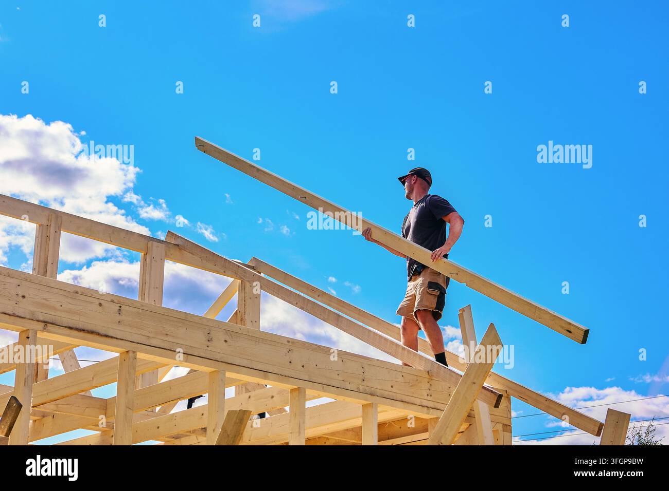 Fissando una trave sulla trave di colmo e sulla piastra murale, un giovane costruttore posiziona un elemento del tetto per l'assemblaggio della struttura in legno. Foto Stock