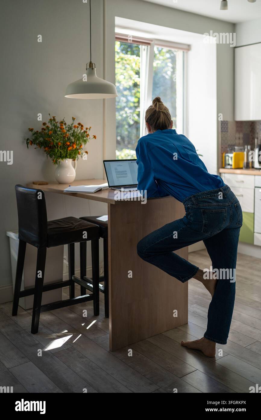 Donna lavora su un laptop da casa in piedi in una posizione di lavoro malsana a un tavolo alto della cucina. Foto Stock