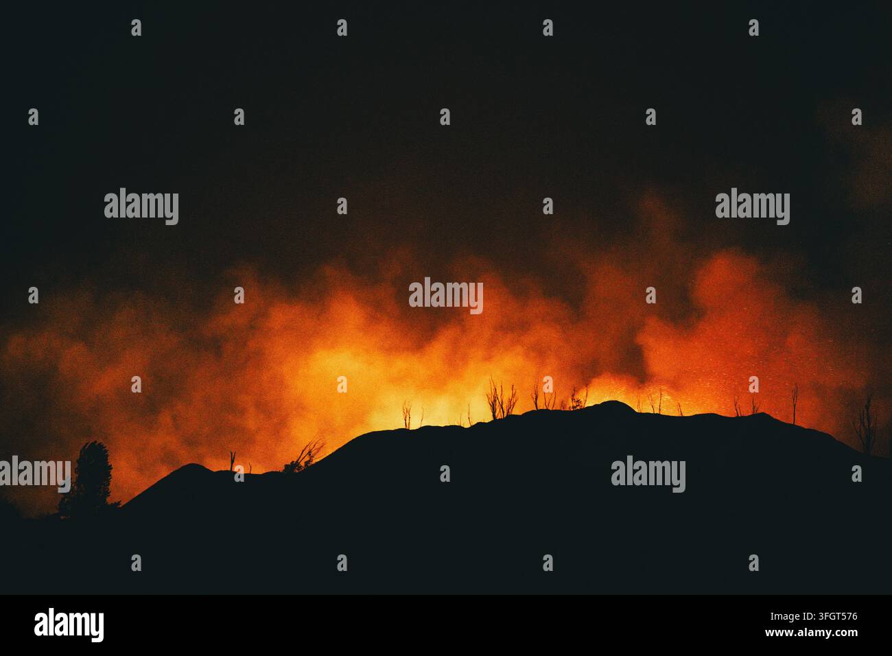 Cresta montuosa in silhouette e alberi bruciati delineati contro fumo e fuoco arancione brillante di notte. Messa a fuoco selettiva. Foto Stock