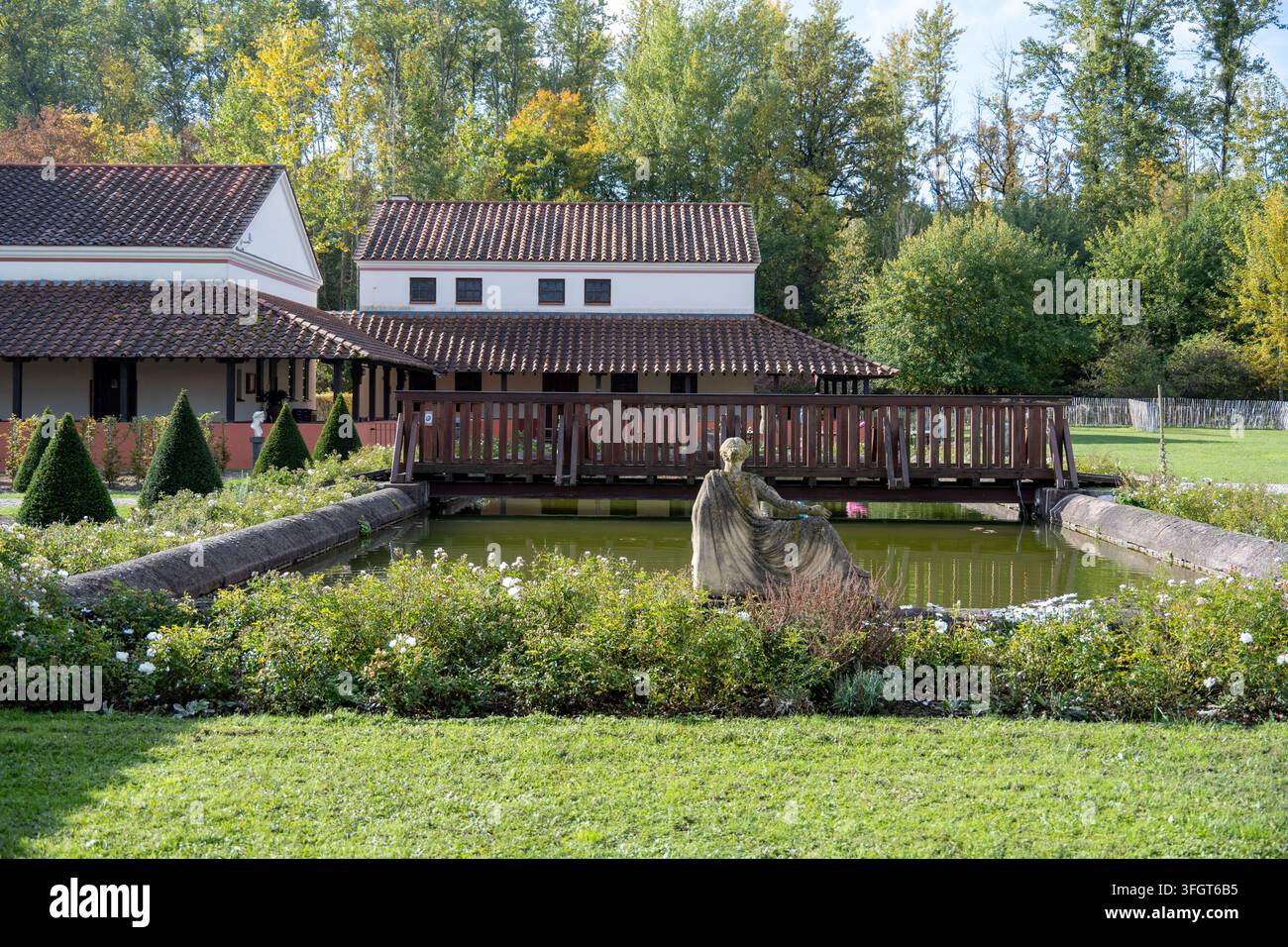 Giardino romano ricostruito con uno stagno rettangolare, ponte di legno, statua classica, arbusti potati, fiori e edifici di villa in Saarland. Foto Stock