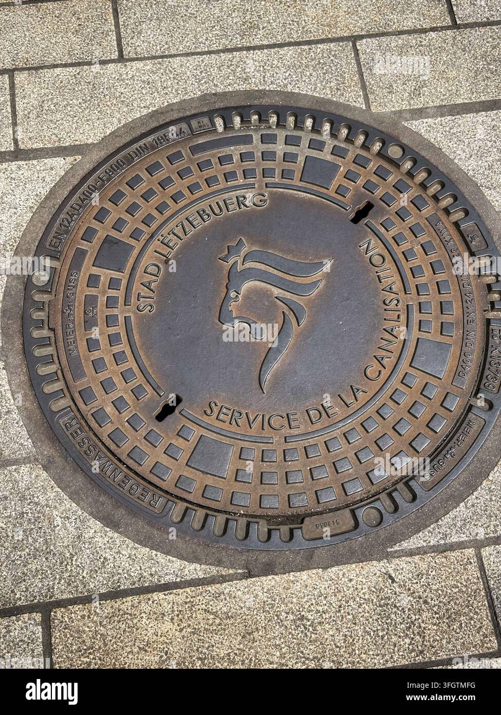 Copertino di grondaia in ghisa con testo della città ed emblema del leone su pavimentazione in pietra. Primo piano dettagliato di una copertura utilitaria rotonda in metallo incastrata in un marciapiede in pietra Foto Stock