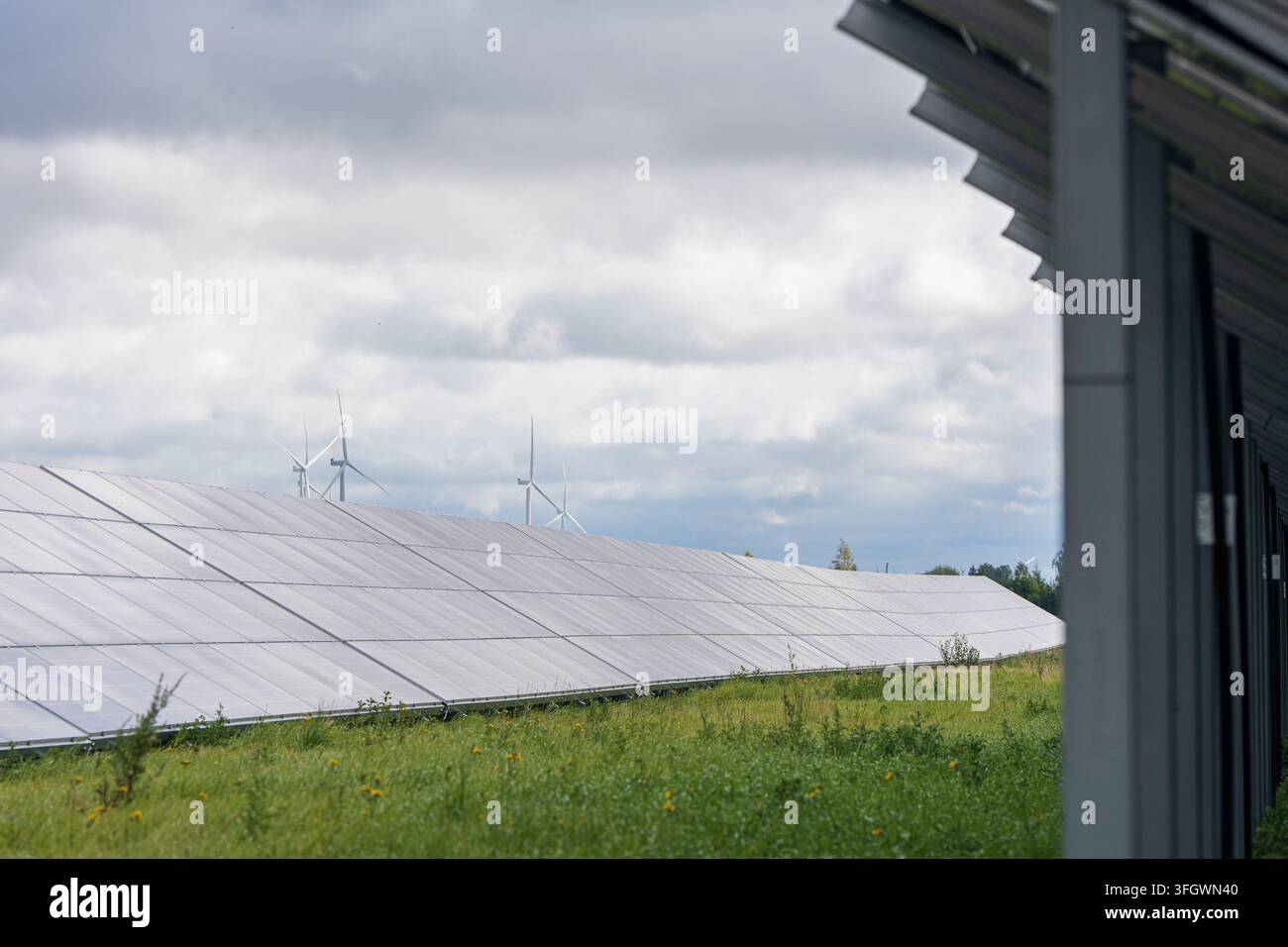Array di pannelli solari e turbine eoliche in un paesaggio rurale verde incorniciato dal bordo dell'edificio sotto un cielo nuvoloso, energia rinnovabile ibrida e produzione di energia sostenibile Foto Stock