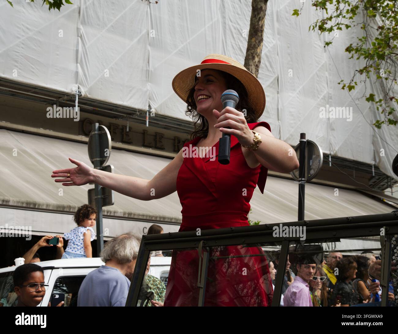 Una donna in un vestito rosso vintage e cappello di paglia che parla in un microfono da un veicolo aperto durante una parata di strada a Parigi, Francia. Foto Stock