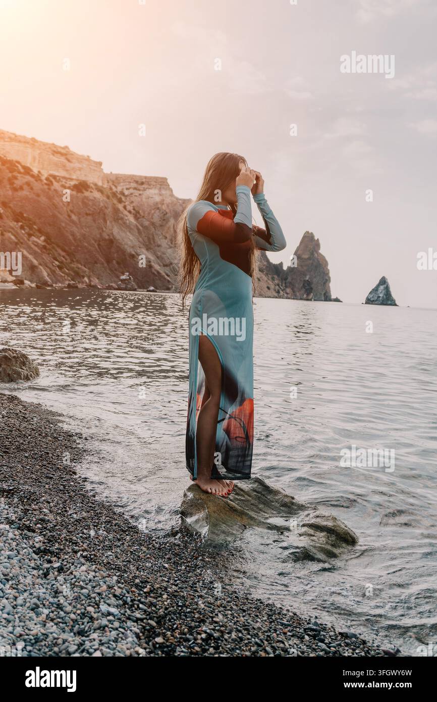 Woman Dress Beach Seascape - Una donna con un abito lungo si erge su una spiaggia rocciosa che si affaccia sul mare. Foto Stock