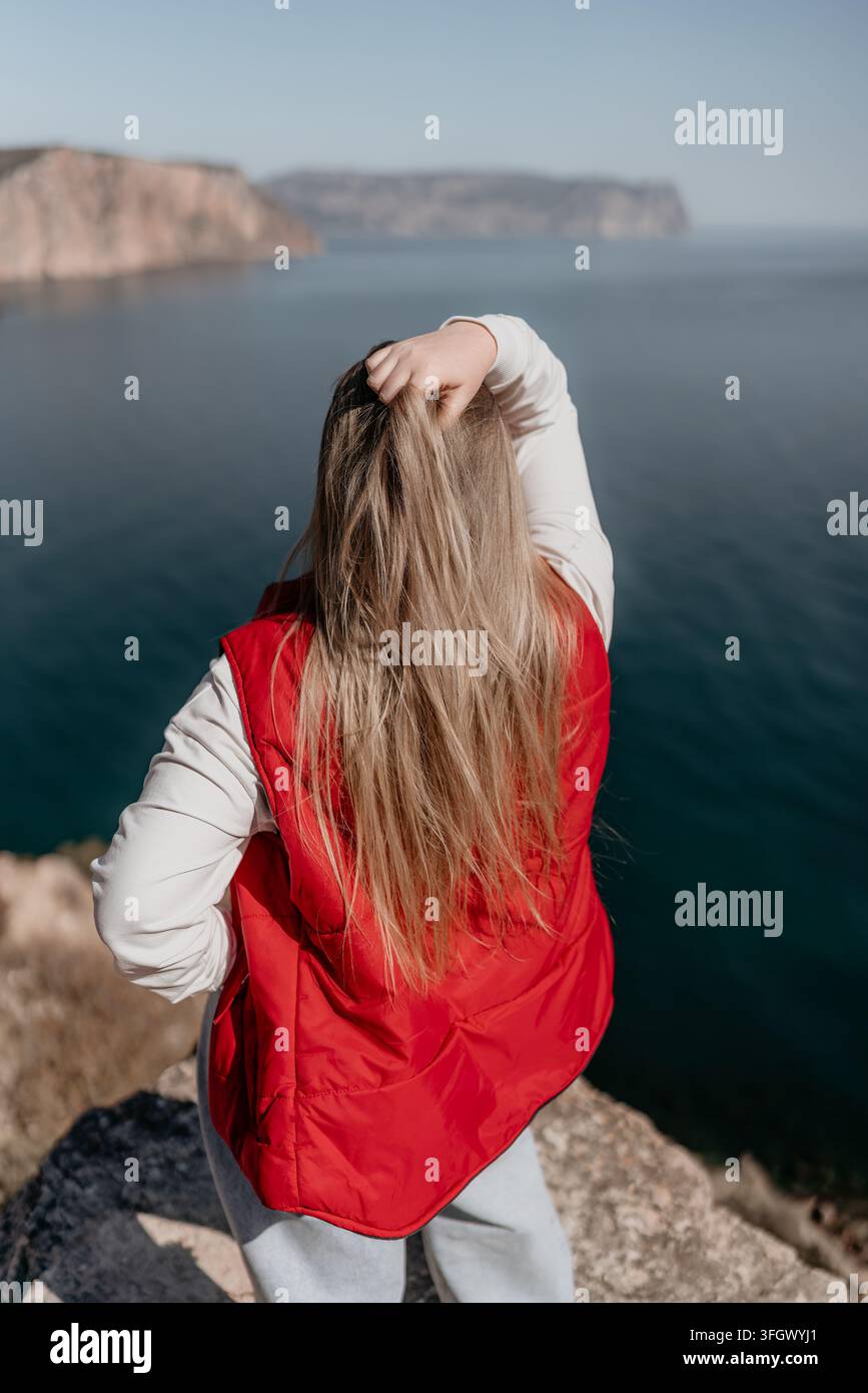 Donna viaggio costiero vista posteriore di una donna con lunghi capelli biondi in un giubbotto rosso che guarda il mare da una scogliera Foto Stock