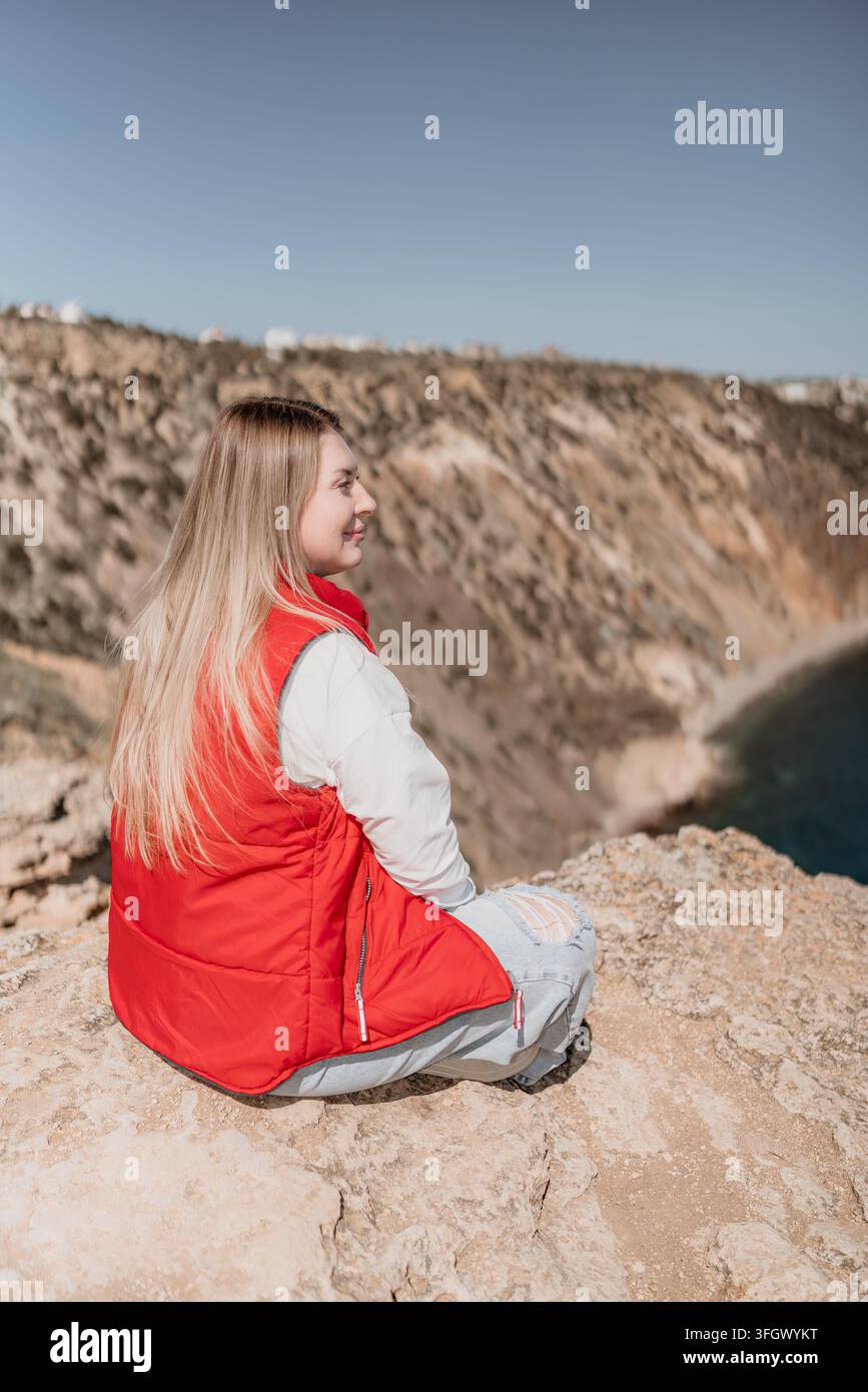 Donna seduta sul bordo roccioso di una scogliera che si affaccia sull'oceano, godendosi il paesaggio costiero, la natura e la libertà durante il viaggio Foto Stock