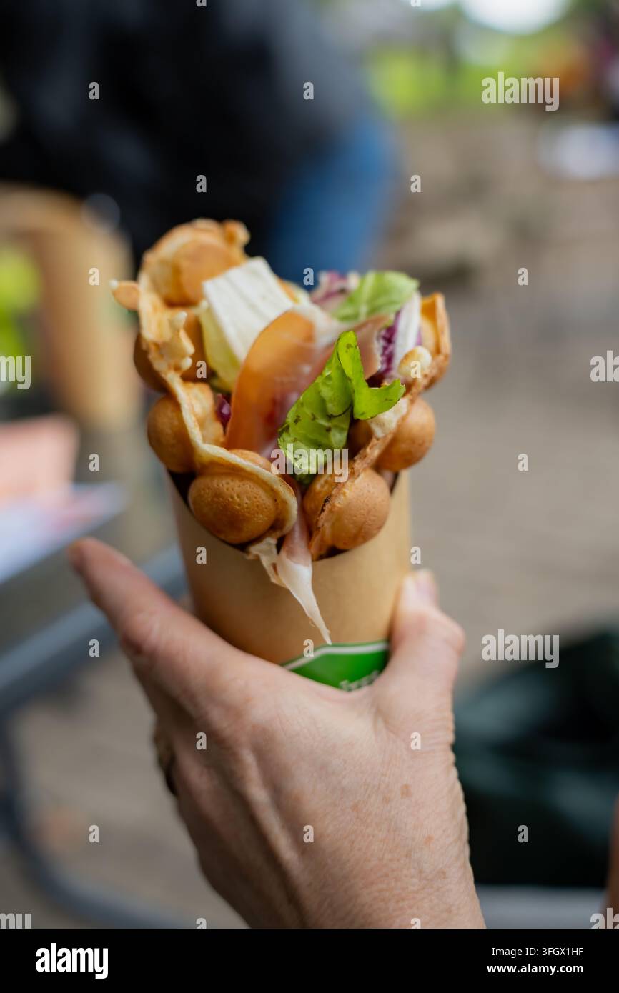 Mano che presenta un bubble waffle farcito con formaggio visibile e insalata a foglia, rappresentante snack salati portatili e pasti confezionati per il cibo da strada Foto Stock