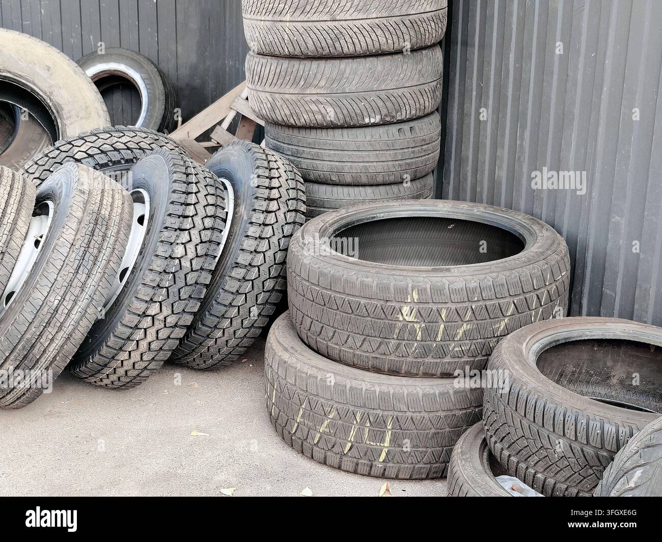 Gomme usate accatastate e inclinate esposte all'aperto contro un muro ondulato scuro, che mostrano diversi modelli di battistrada, dimensioni, superfici consumate e gravità Foto Stock