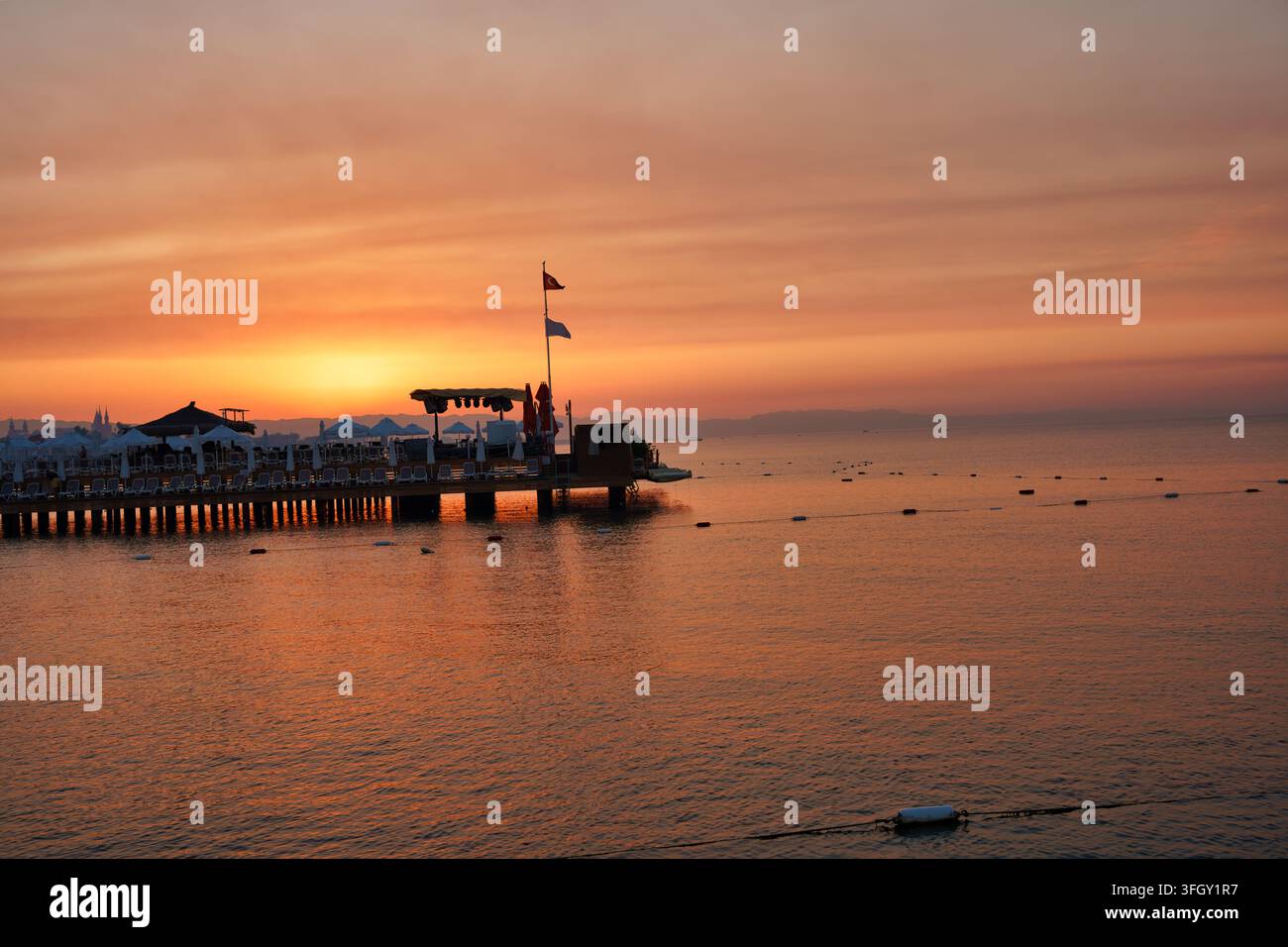 Silhouette di un molo di legno del resort con bandiere e ombrelloni che si estendono nelle acque calme dell'oceano durante un tramonto o alba dorato e arancione. Bella costa Foto Stock