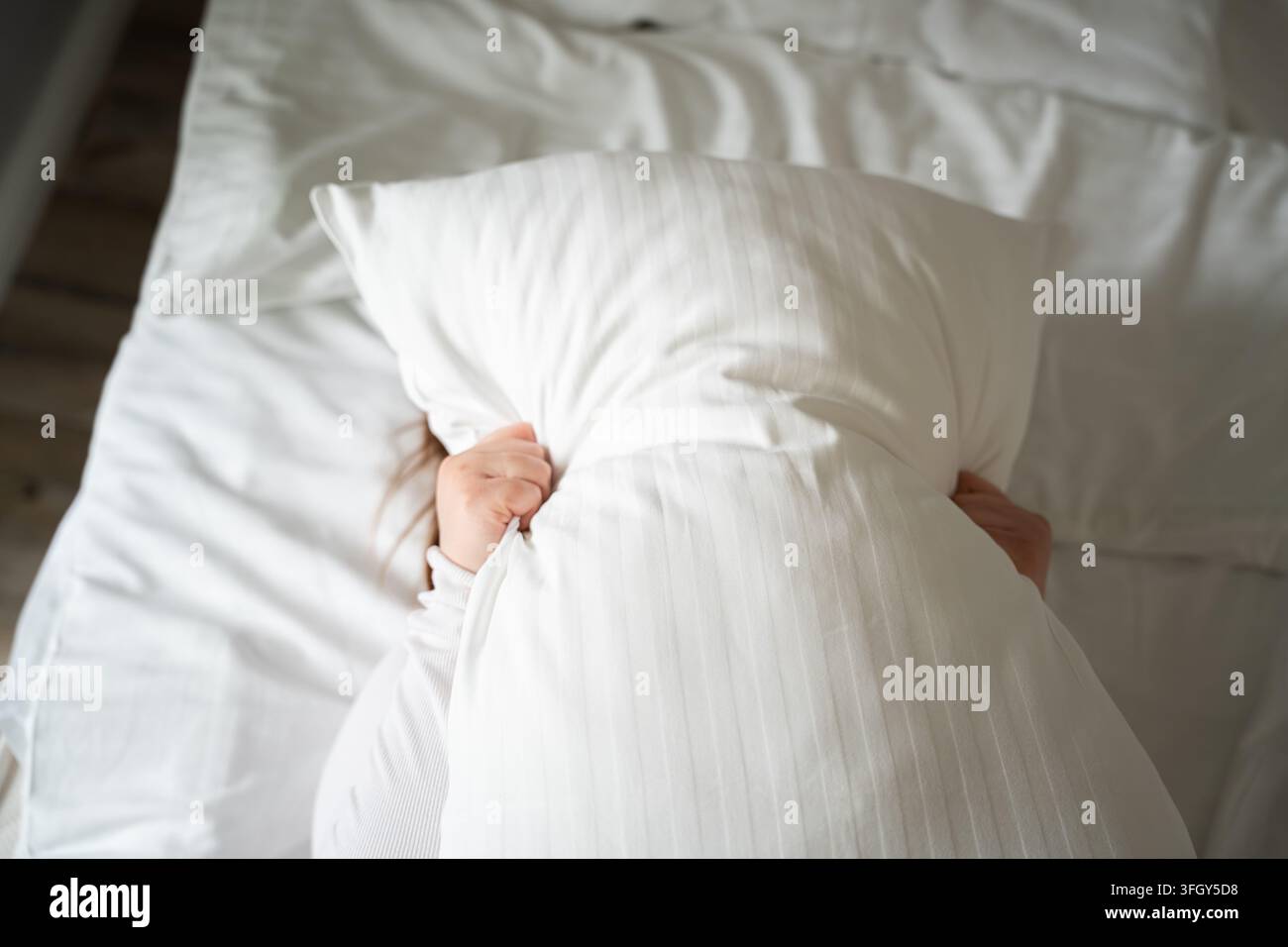 Adulto a riposo a letto mentre tiene un cuscino bianco sul viso in una camera da letto luminosa. Disturbo del sonno, sollievo dallo stress, spazio personale e blocco del rumore Foto Stock
