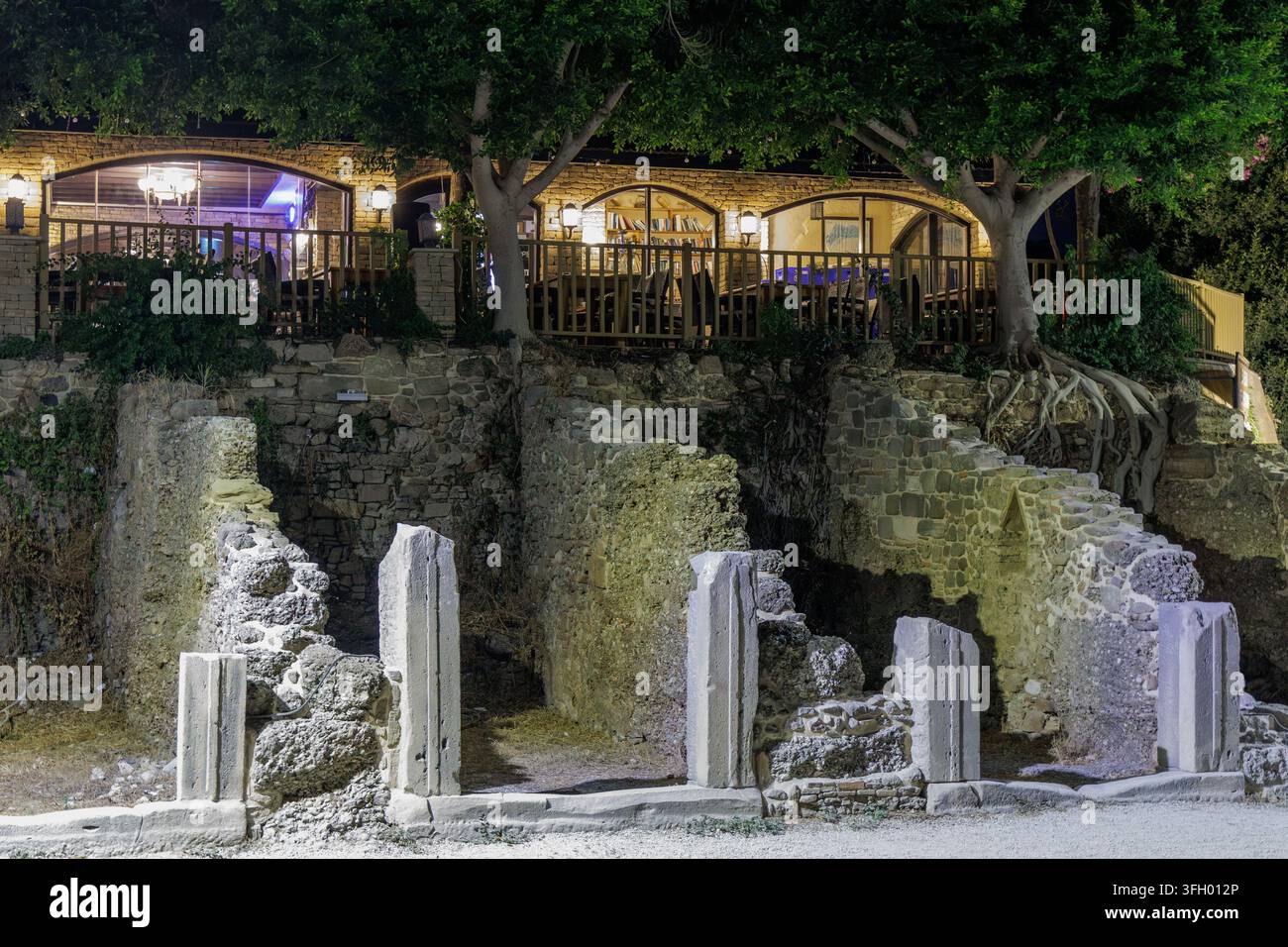Bellissimo contrasto tra rovine storiche in pietra e colonne con un moderno edificio accogliente di un caffè all'aperto sotto alberi verdi nella vecchia città di Side di notte Foto Stock