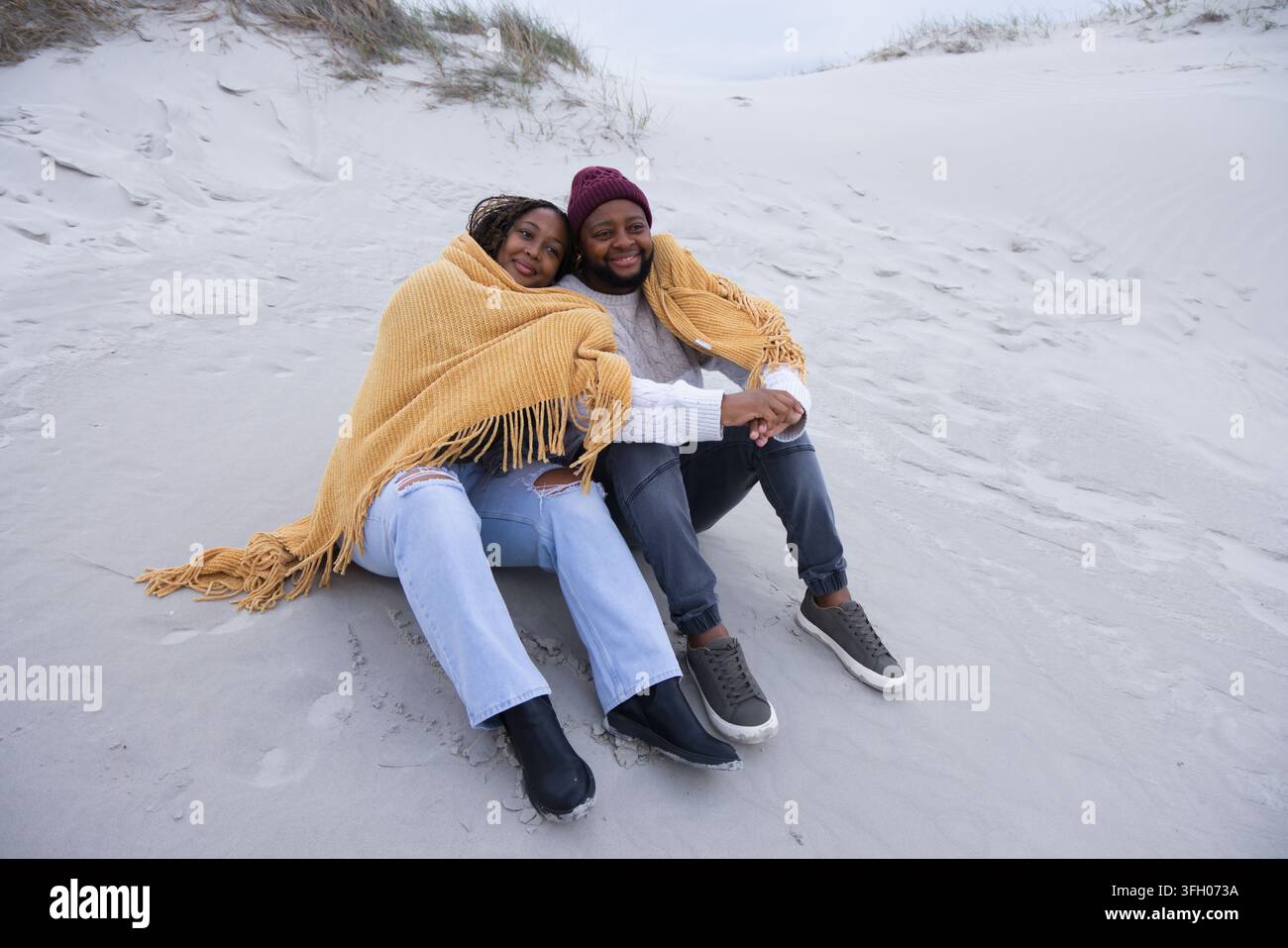 Coppia seduta avvolta in una coperta giallo senape sulle dune di sabbia, con un berretto marrone Foto Stock