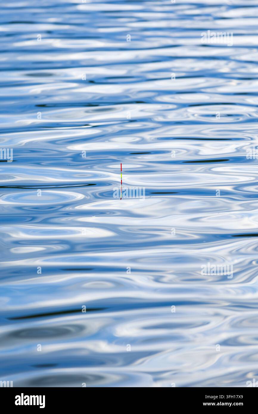 Un galleggiante da pesca dai colori vivaci galleggia sulla superficie increspata di un lago blu tranquillo, creando una scena pacifica di pesca dolce silenziosa durante un sole Foto Stock