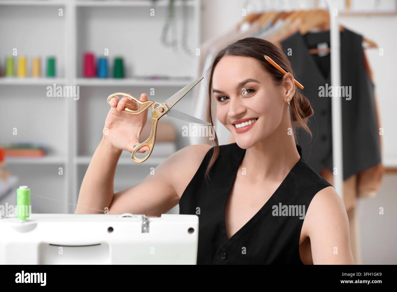 Azienda di design della moda e sartoria, sarta sorridente con le forbici in uno studio di cucito creativo con espositori di abiti e bobine di filo Foto Stock