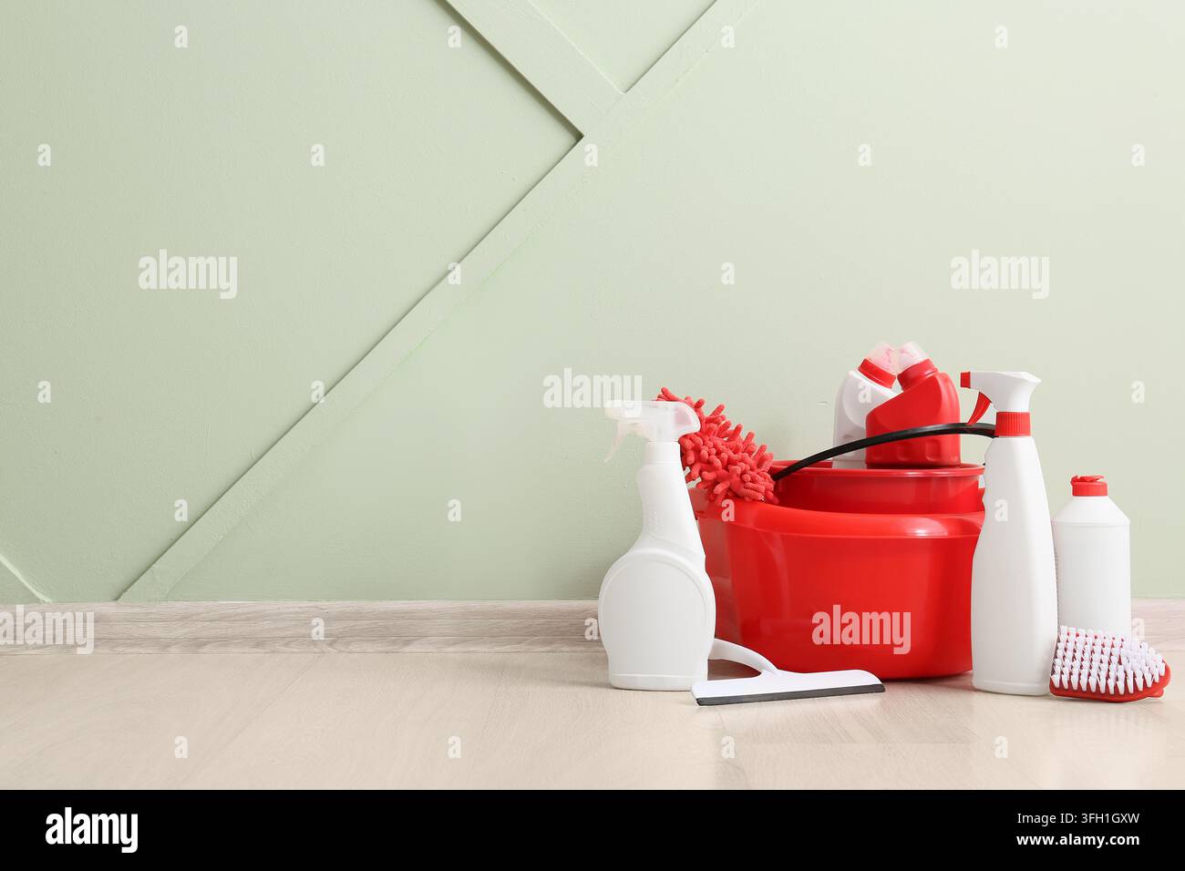 Servizio di pulizia della casa e pulizia di primavera, set di utensili professionali e detergenti in un secchio rosso su un pavimento di legno contro un muro verde chiaro Foto Stock